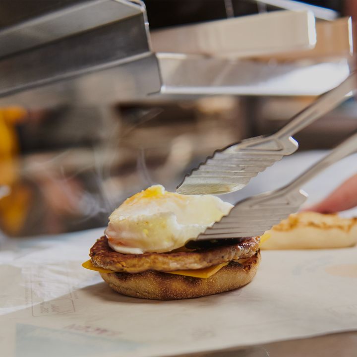 Tongs place a cooked egg on a sausage patty and cheese, all on an English muffin, in a busy fast-food kitchen.