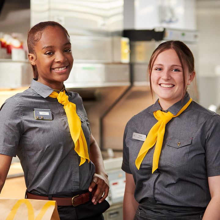 Two employees smiling in gray uniforms with yellow scarves stand in a fast-food restaurant.