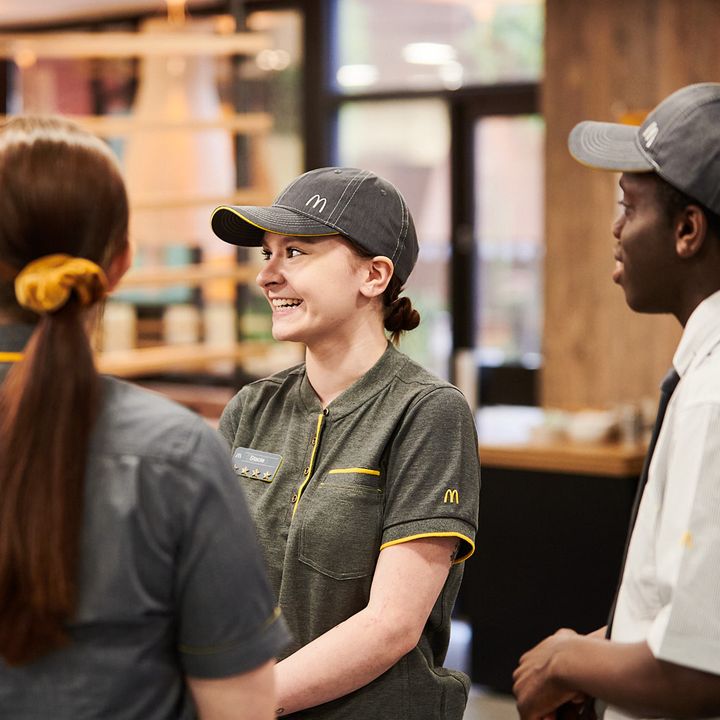 Three employees in a fast-food restaurant smiling, wearing gray polo shirts and black caps.