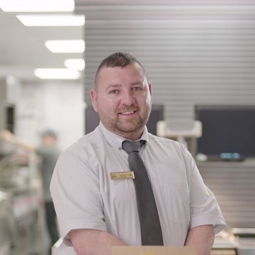 A man smiling wearing a white shirt and grey tie, stands in a fast-food kitchen.