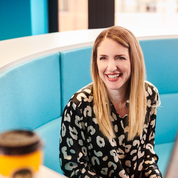 A woman smiling, sitting on a blue padded sofa in an office setting.