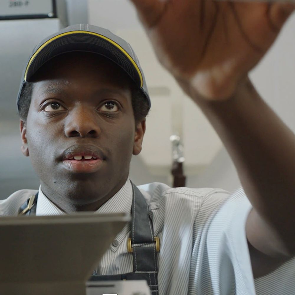 A man in grey uniform and a cap reaching up to grab something, in a fast-food kitchen.