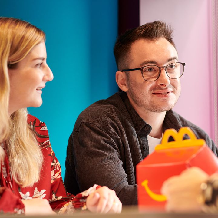 Two individuals at a table, both smiling, in an office setting. There is food packaging on the table.