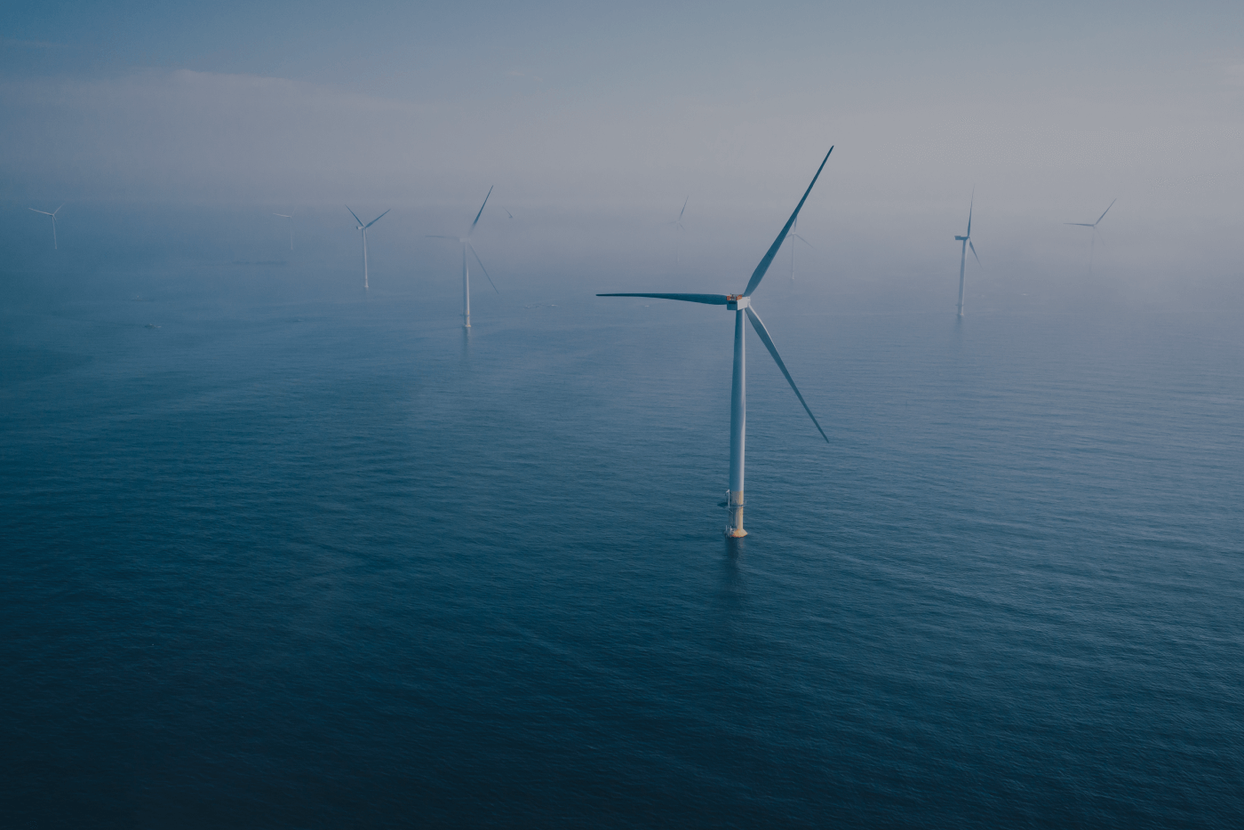 Who we are_Corporate Responsibility.Multiple offshore wind turbines standing in calm ocean waters under a hazy sky