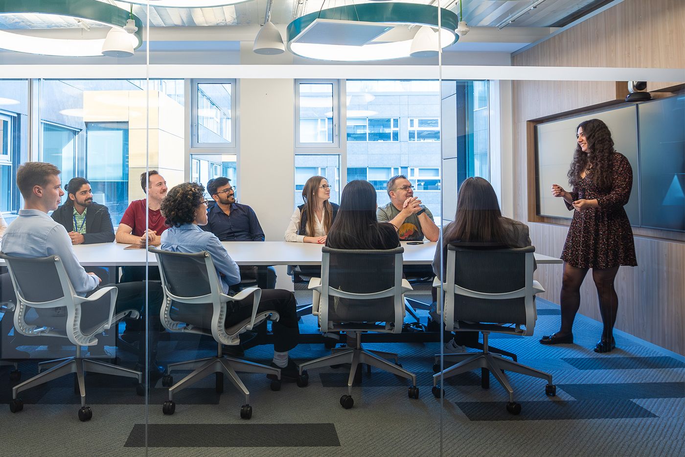 Life at MSCI_Learning.A person standing and presenting to a group of colleagues seated around a conference table in a modern glass-walled meeting room