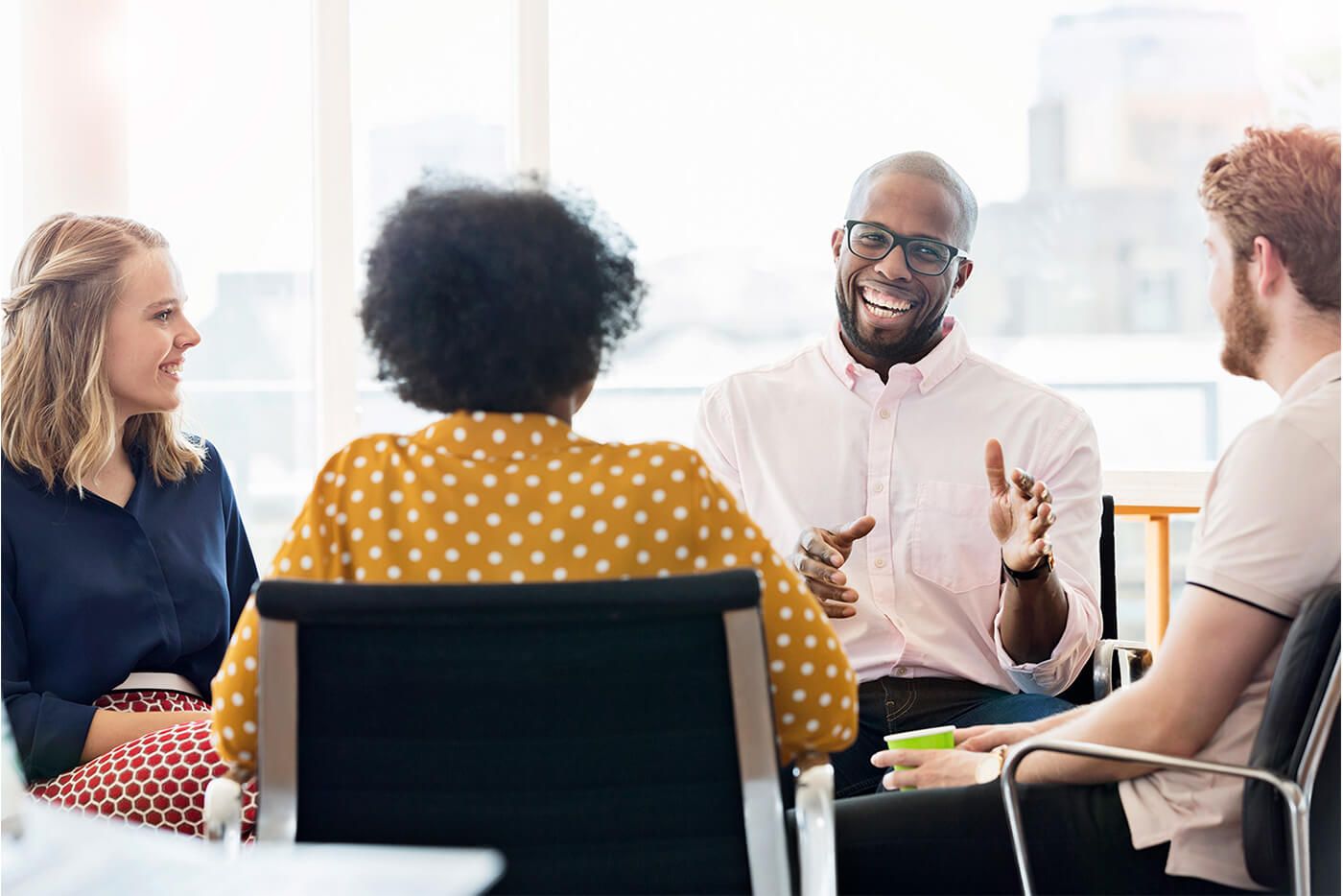 Who we are_Culture.Four people seated in a bright office, engaged in a lively group conversation, with one person gesturing while speaking and others smiling and listening.