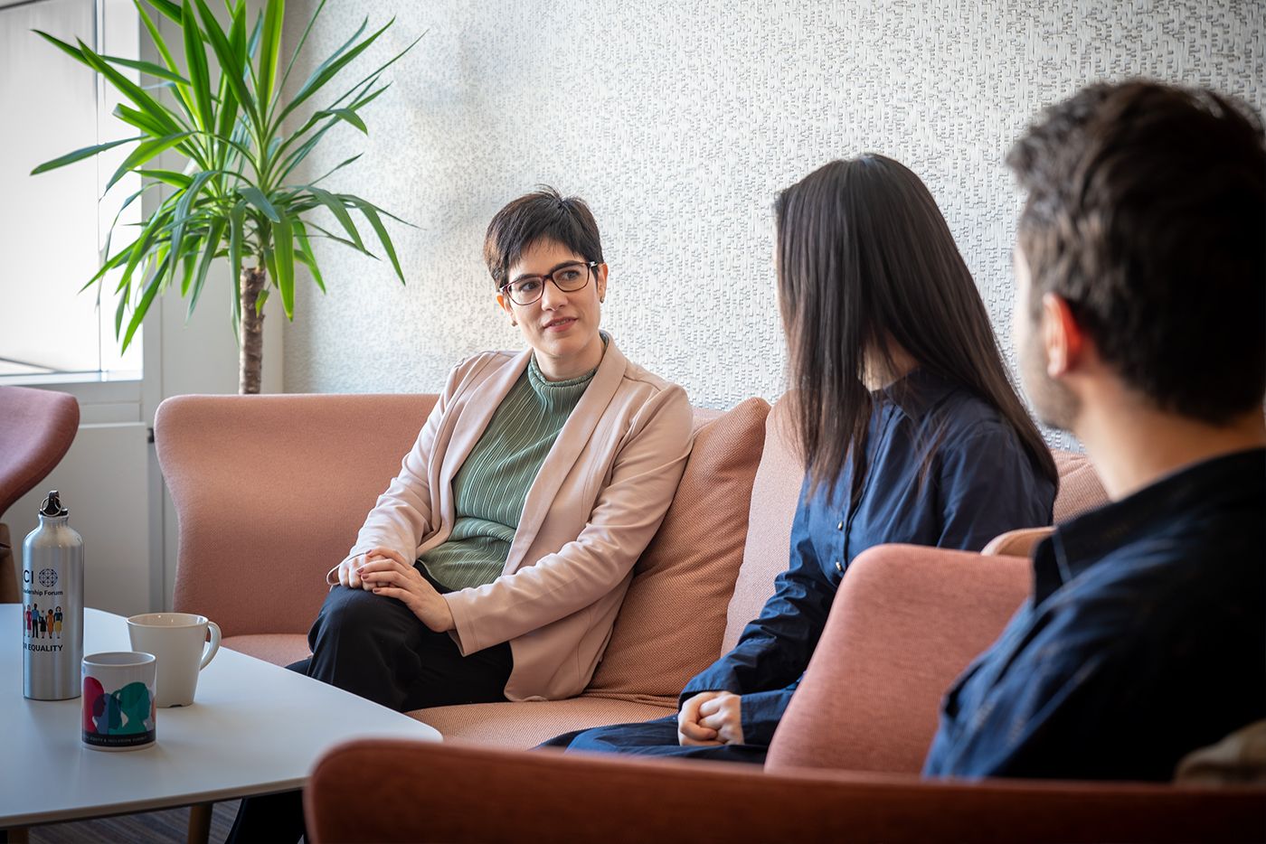 Life at MSCI_Development. Three colleagues sitting on a sofa in a casual office lounge, having a relaxed conversation near a coffee table and potted plant.