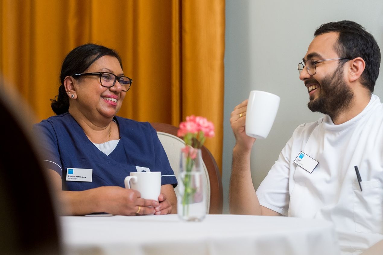 Two Bupa Care Services colleagues in uniform sitting at a table, talking over a drink during a break in a calm, welcoming workplace setting.
