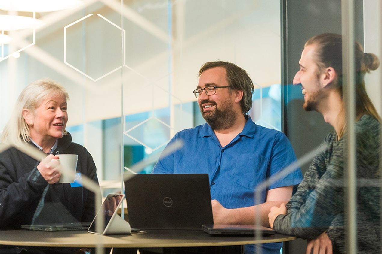 Three people seated around a round table in a glass-walled meeting room, with laptops open and a mug on the table. Geometric patterns are visible on the glass walls, and the office environment is visible in the background