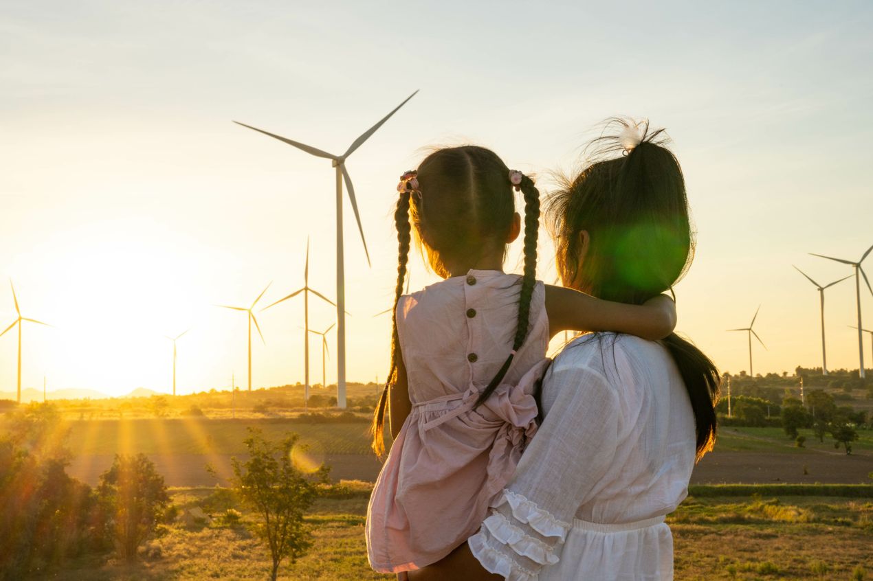 Person holding a child while looking across a landscape of wind turbines at sunset, with open fields, trees, and multiple wind turbines visible against the evening sky