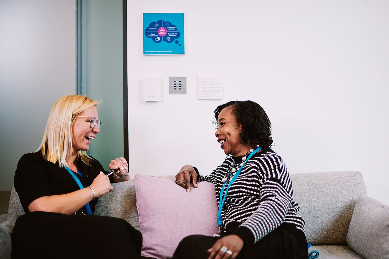 Two colleagues wearing blue lanyards seated on a sofa in an office setting, engaged in conversation. A cushion is placed between them, and a wall poster is visible in the background